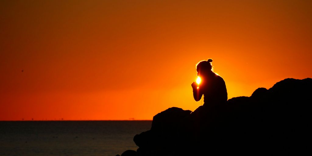 silhouette of person sitting on rock near body of water during sunset